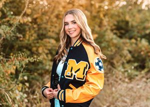 A varsity letterman jacket in black and gold is worn outdoors during autumn with fall foliage in the background.