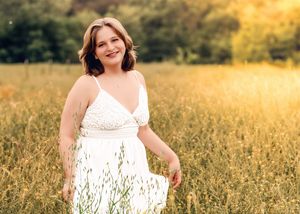 A figure in a flowing white dress stands gracefully in a sunlit meadow of tall grass and wildflowers during golden hour.
