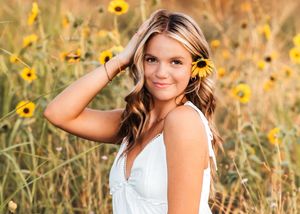A person poses in a sunflower field at golden hour wearing a white dress with flowers in their hair.