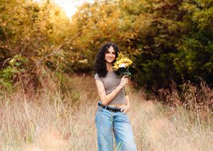 Young woman smiling in a field holding yellow flowers, wearing jeans and a gray top, with autumn trees in background.