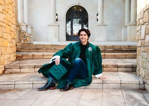 Graduate in green velvet cap and gown sitting on stone steps outside a classical building, smiling.