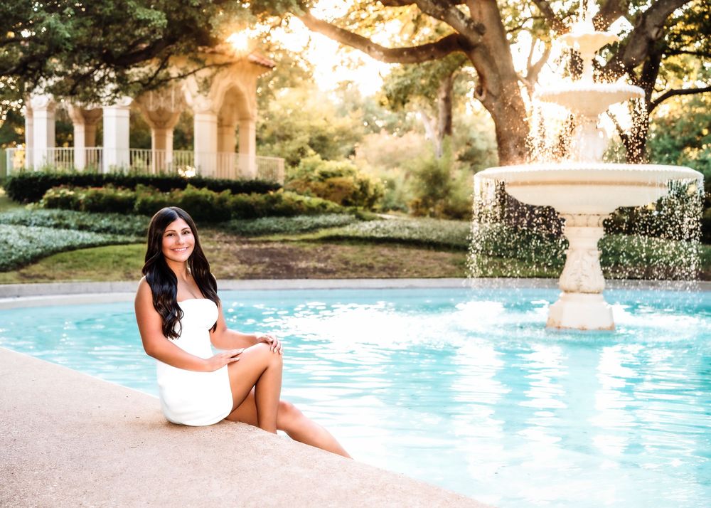 Young woman in white dress sitting poolside near a stone fountain in a lush garden at golden hour.