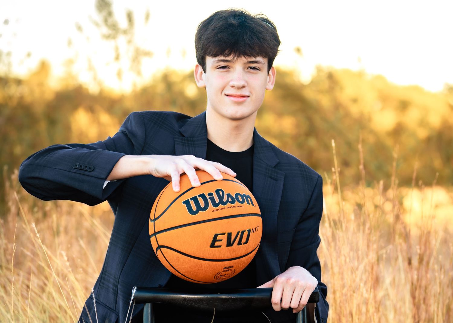 Young man in suit holding Wilson EVO basketball outdoors in golden field during sunset photography session.