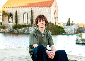 Young man in green striped shirt sitting by a waterfront with a historic stone building in the background.