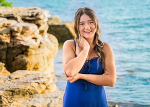 Person in a blue dress poses by rocky coastal cliffs with turquoise ocean waters in the background.