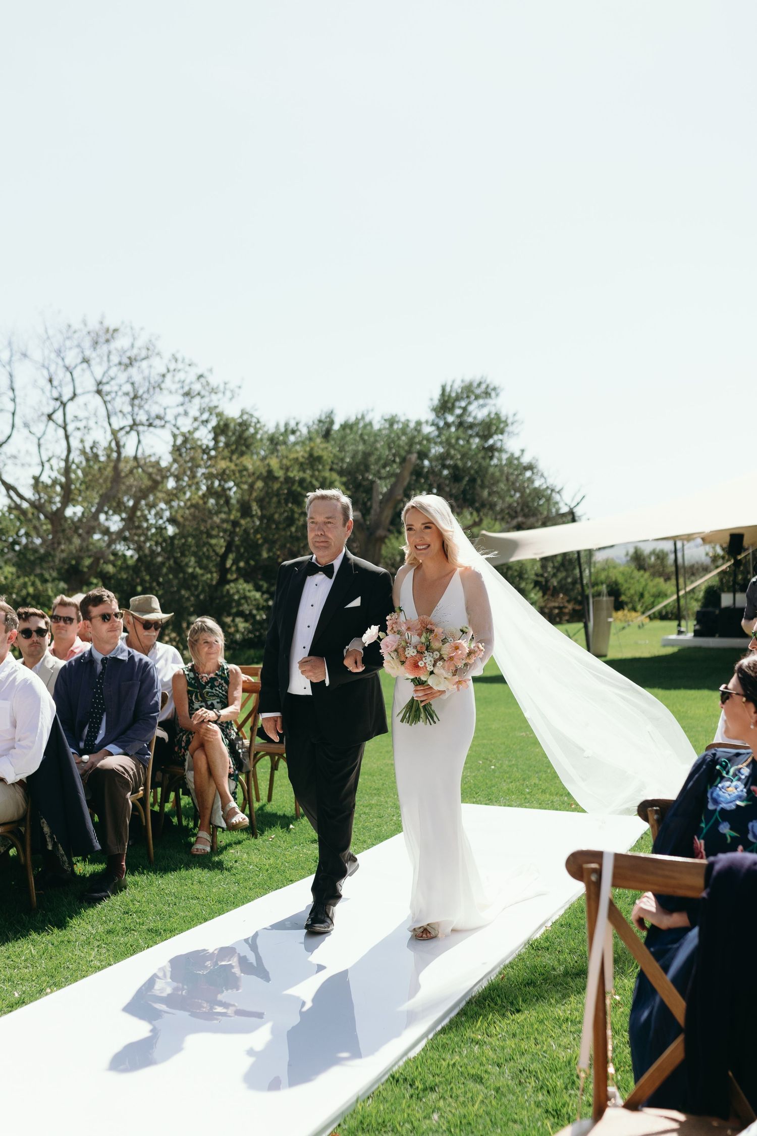 A wedding ceremony takes place outdoors on a sunny day with a white aisle runner and guests seated on either side.