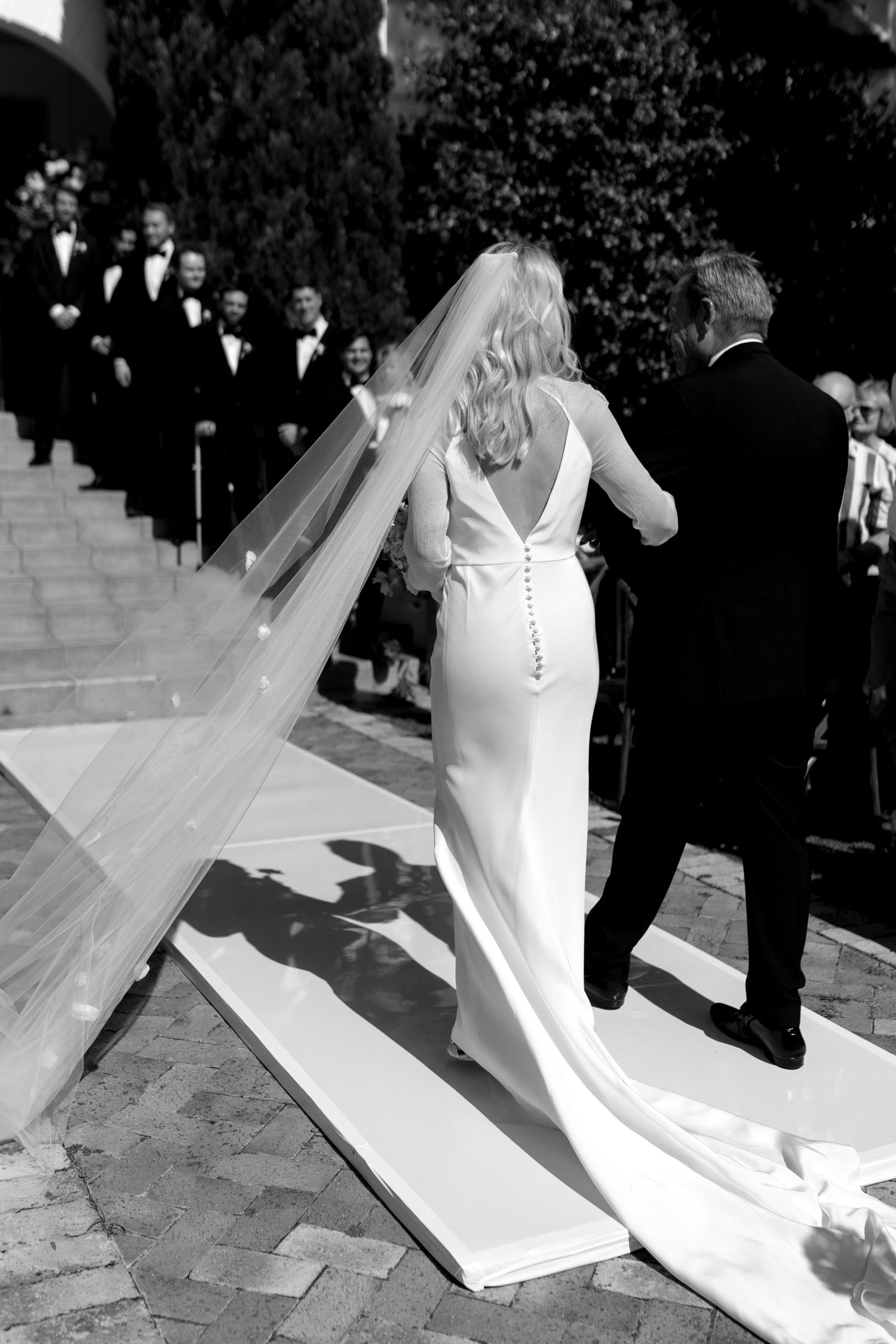 A bride in an elegant white gown walks up stone steps with her flowing veil trailing behind her.