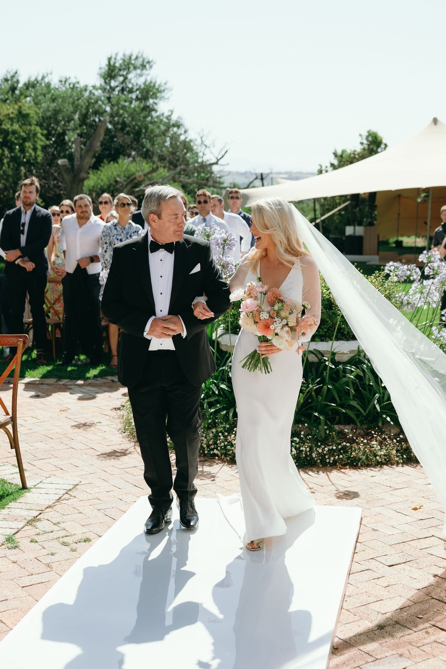 A bride and groom walk down a sunlit stone pathway during their outdoor wedding ceremony.