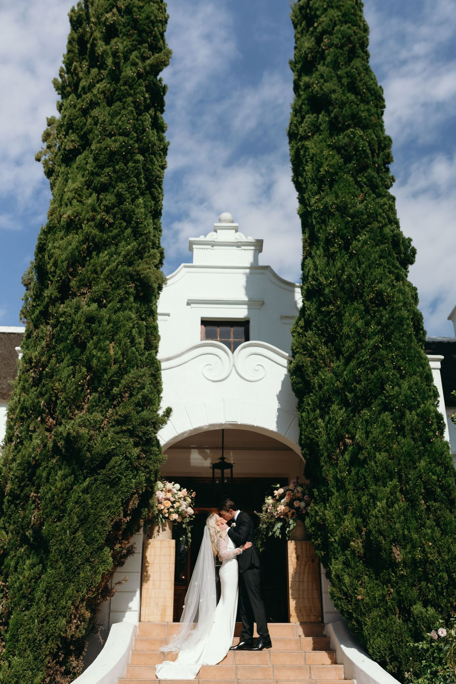 Tall cypress trees line the exterior of a white Spanish colonial style building with arched entryways.