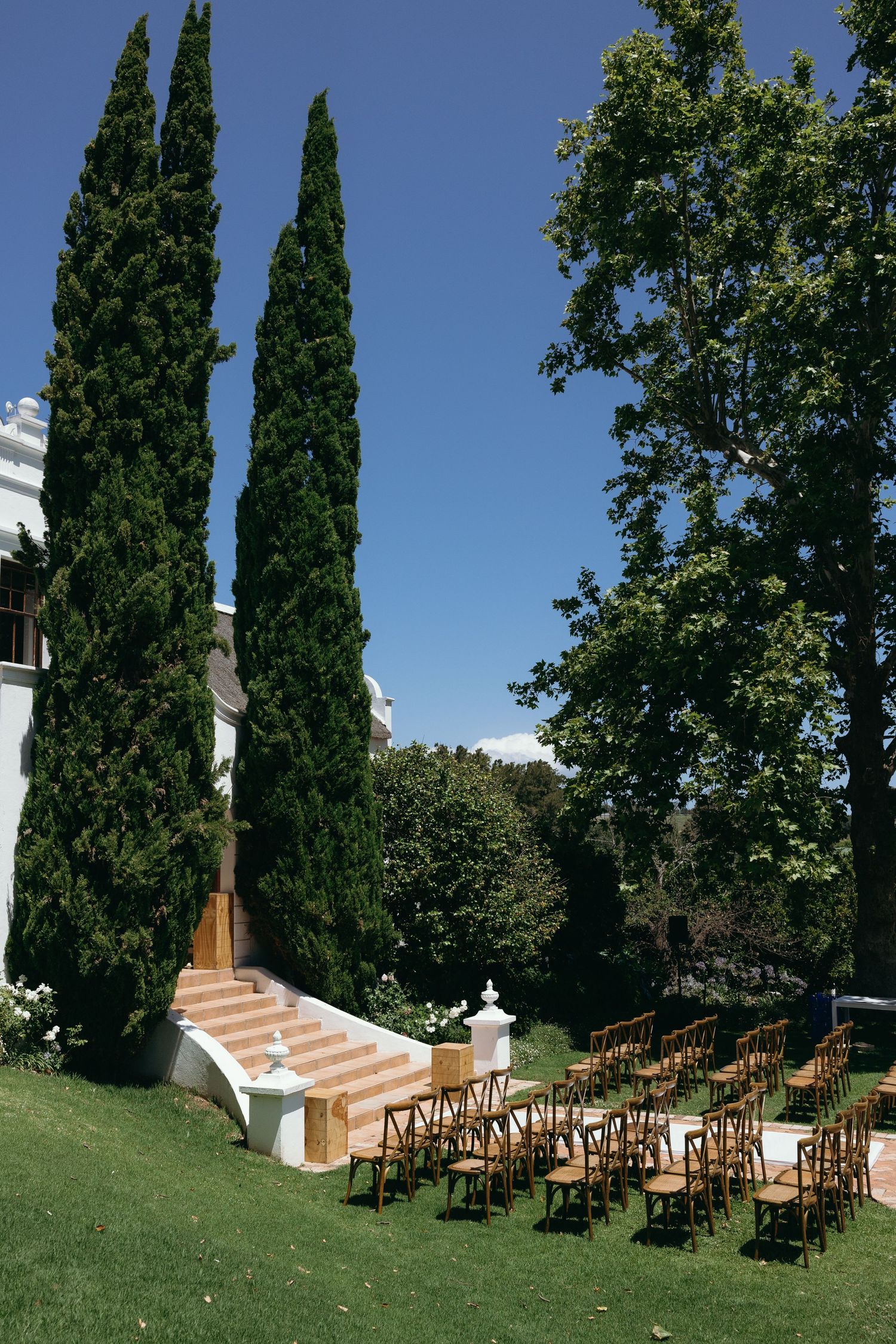 Outdoor wedding ceremony setup with wooden chairs arranged in rows facing a white altar among tall cypress trees.