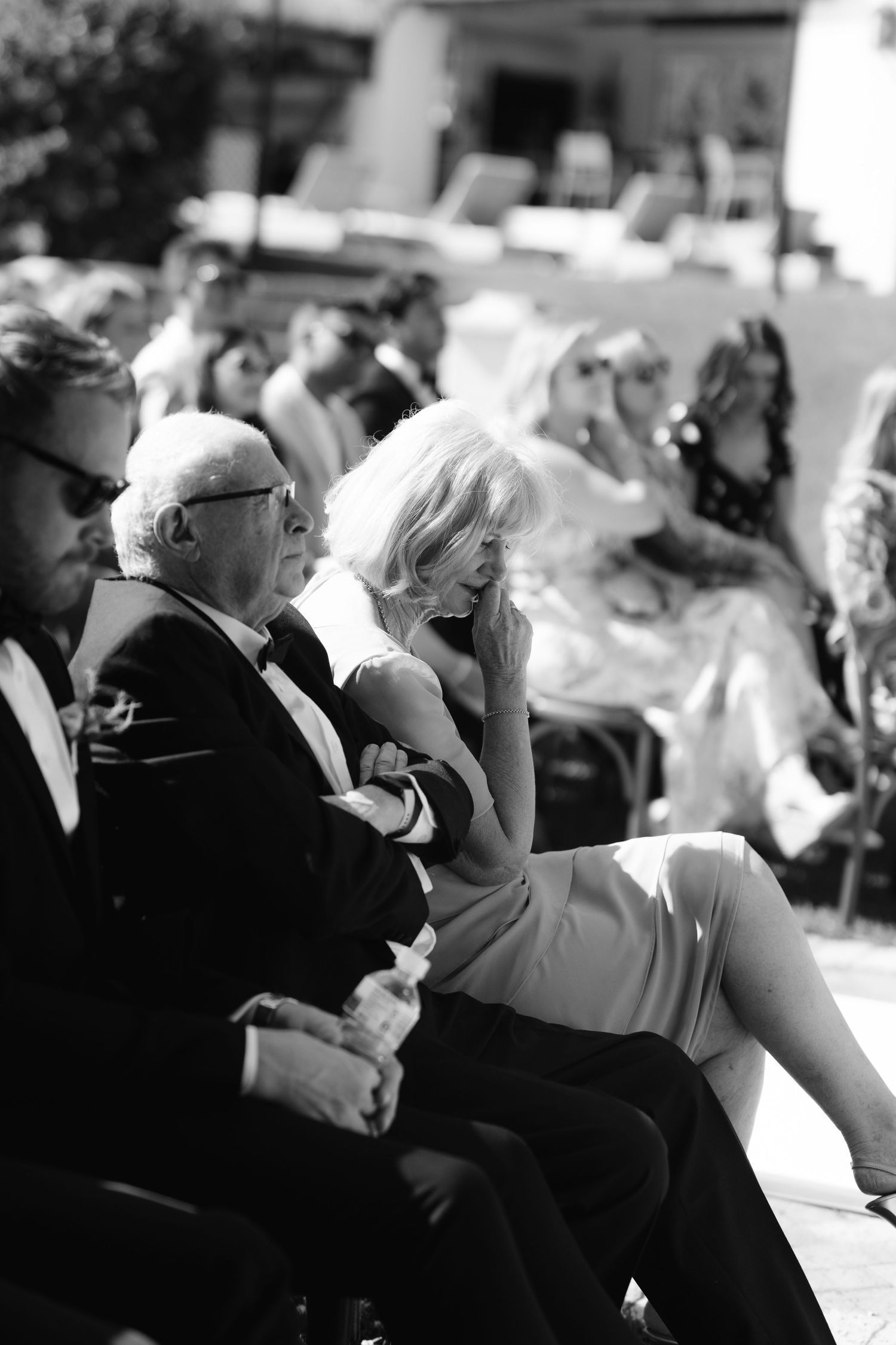 A black and white candid photograph of people seated at an outdoor event, with bright sunlight casting shadows.
