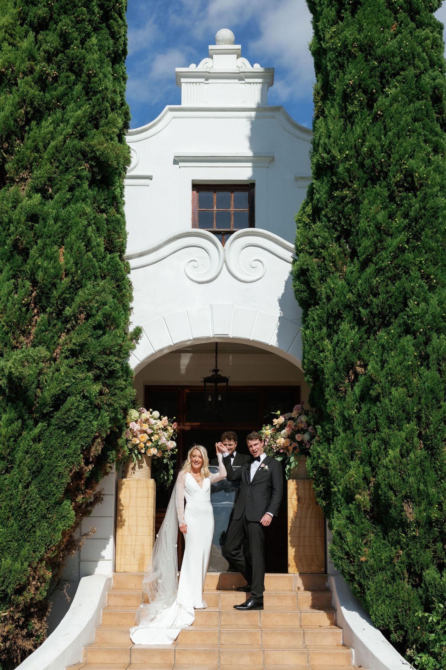 White stucco church entrance with terracotta steps and tall cypress trees framing a wedding ceremony.