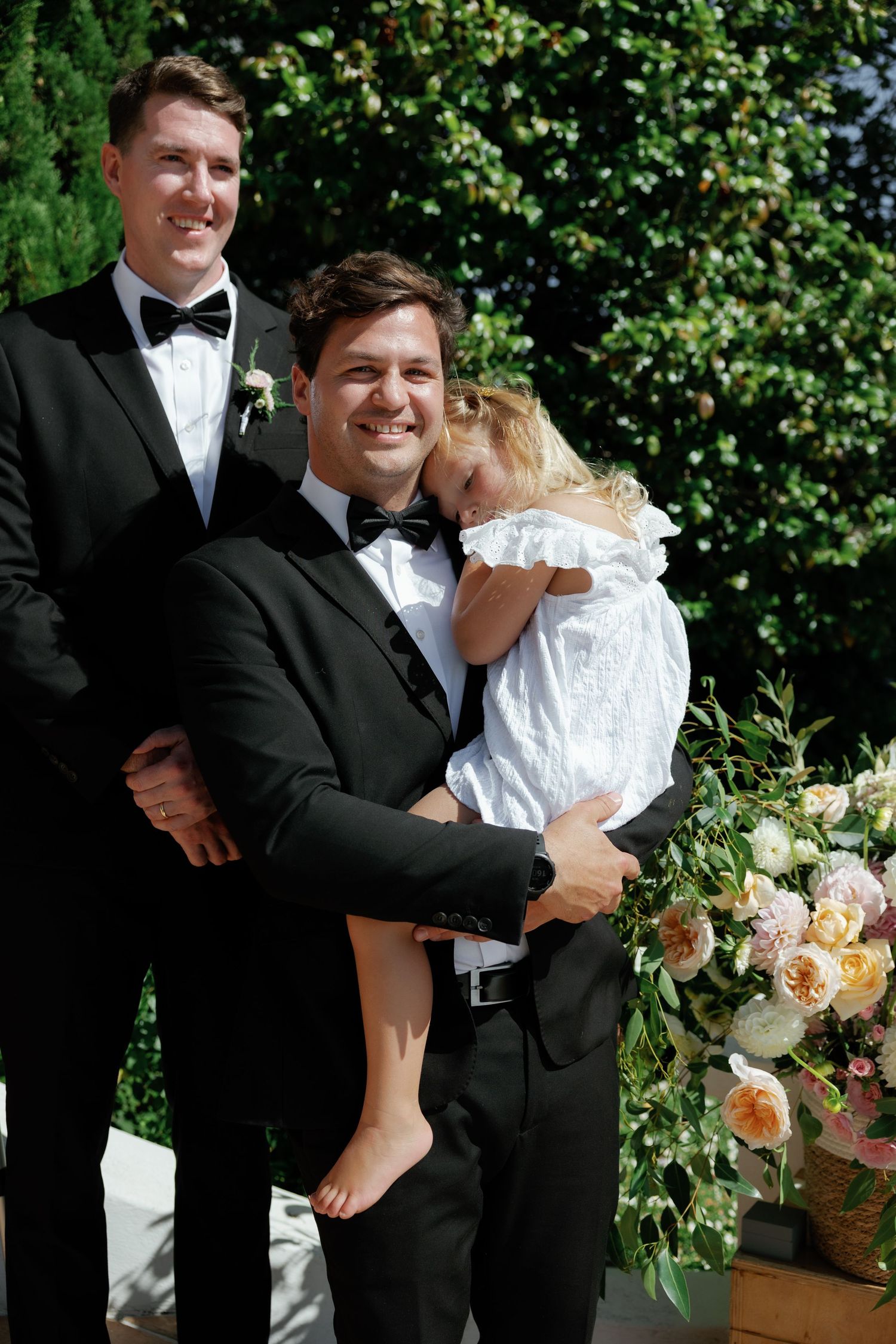A joyful wedding moment captured outdoors with lush greenery and peach-colored floral arrangements in the background.
