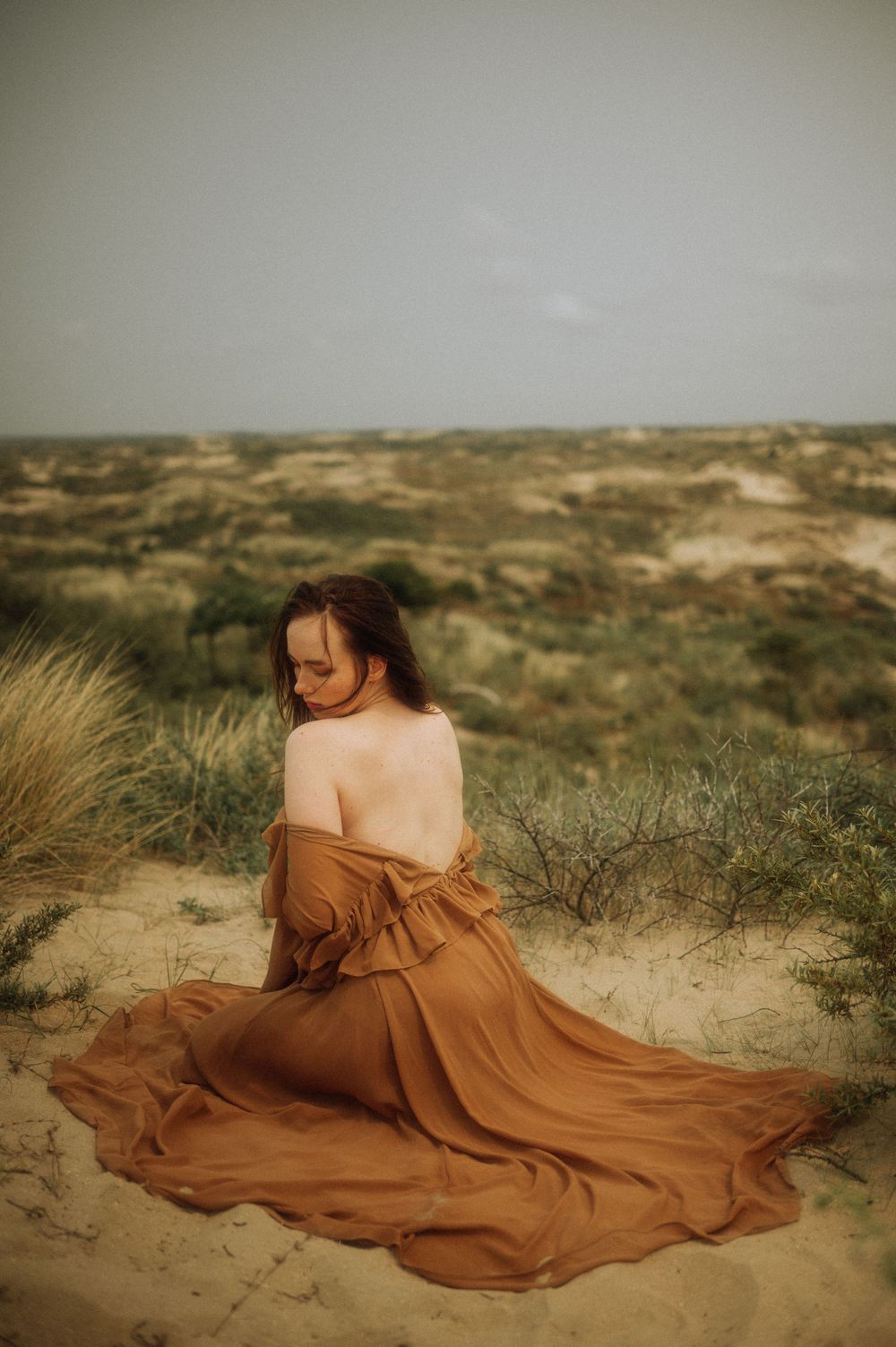 Flowing brown dress billowing in desert landscape against sandy dunes and brush.