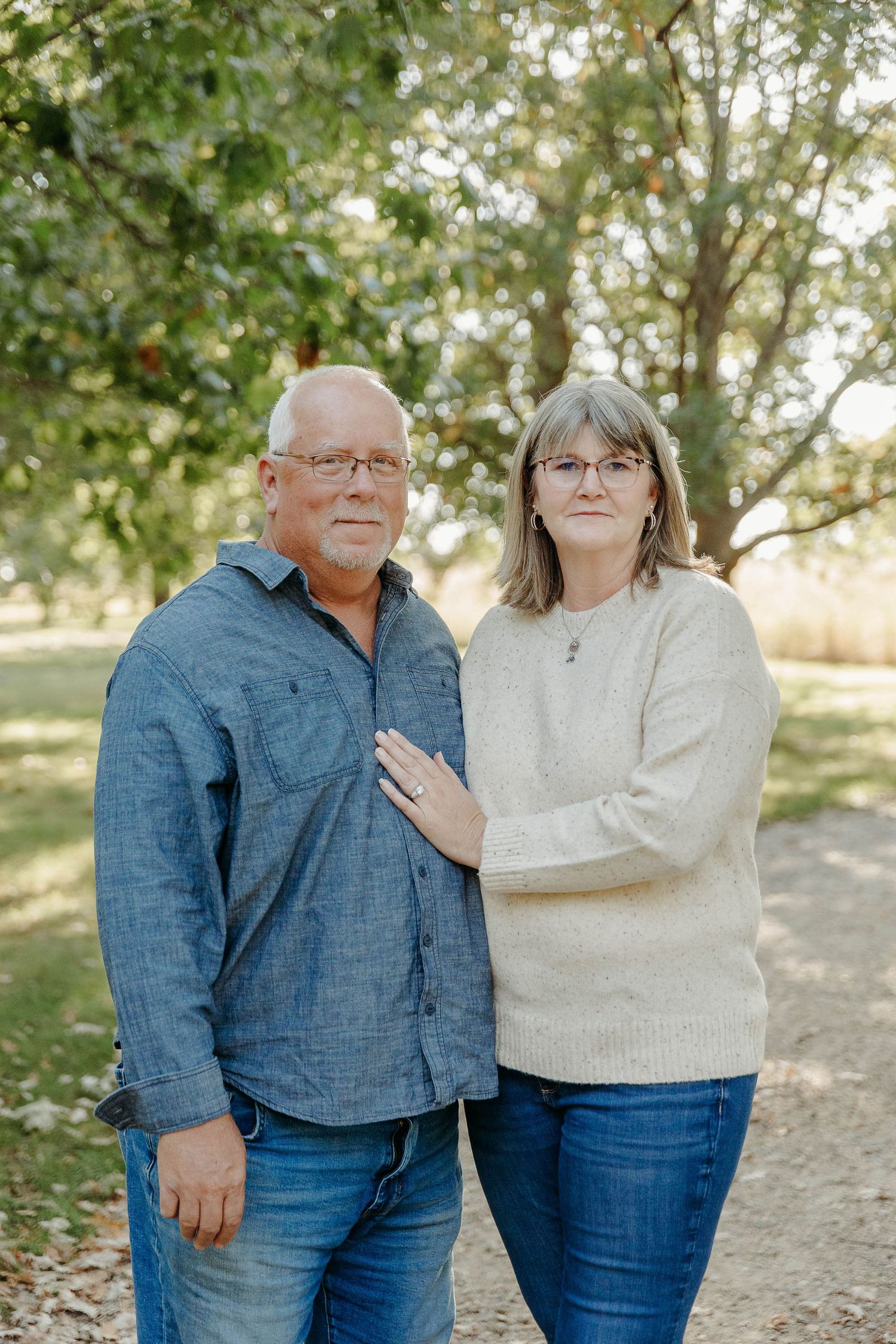 A couple wearing denim and cream colored sweater poses together outdoors among trees on a sunny day.