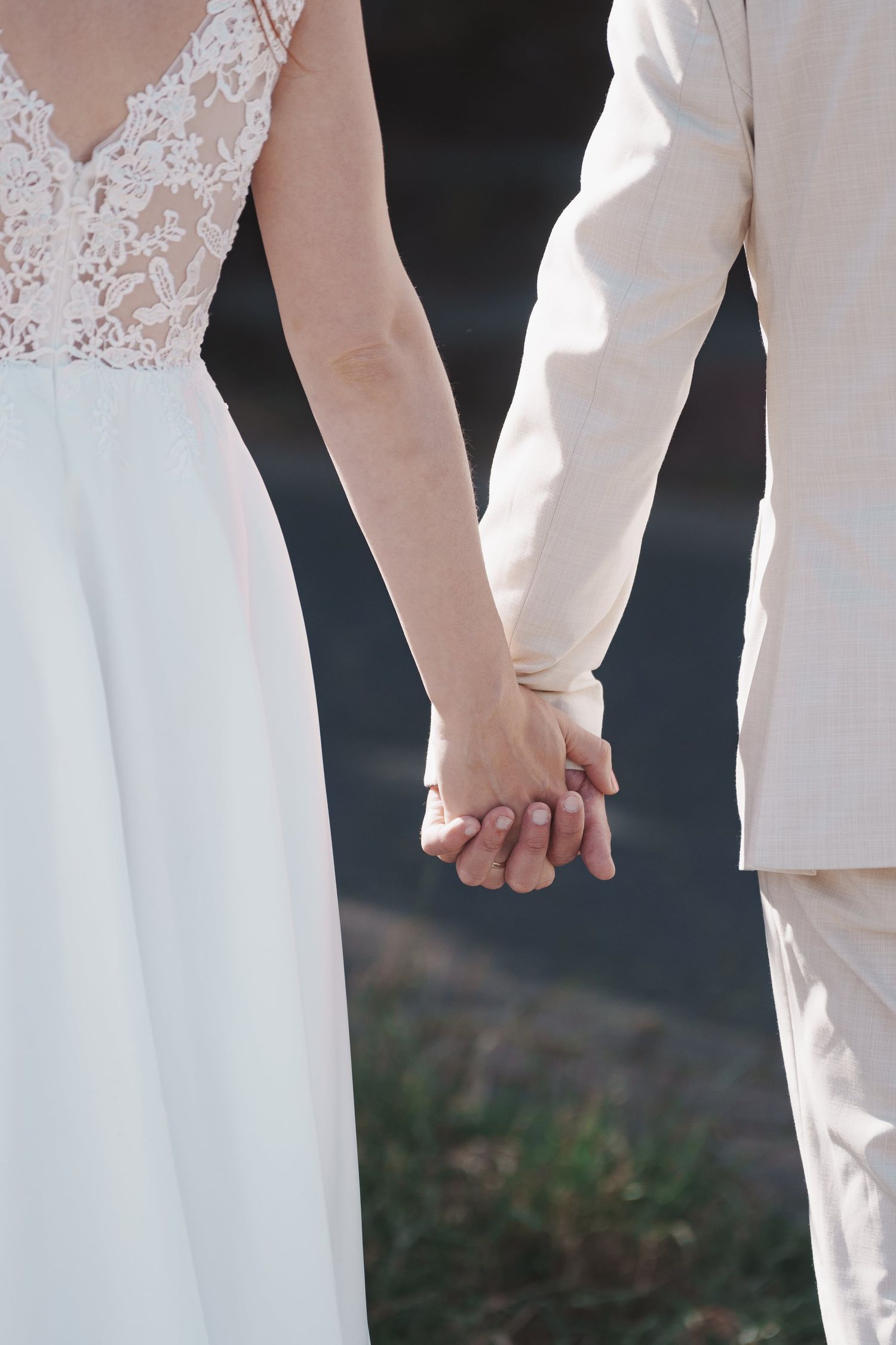 Close-up of bride in lace dress and groom in beige suit tenderly holding hands outdoors.