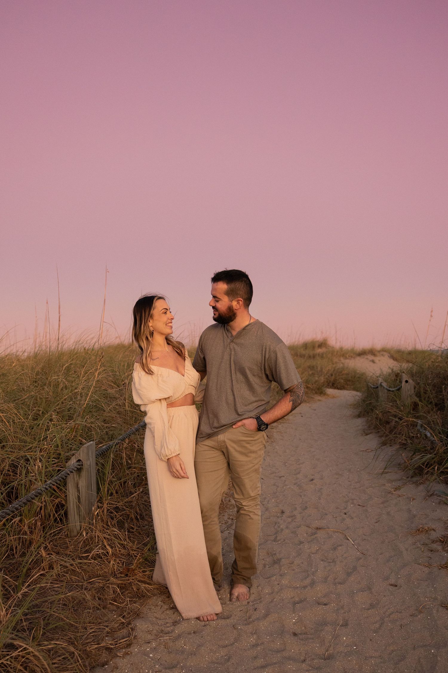 Romantic couple walks sandy beach path lined with sea oats under a beautiful pink sunset sky.