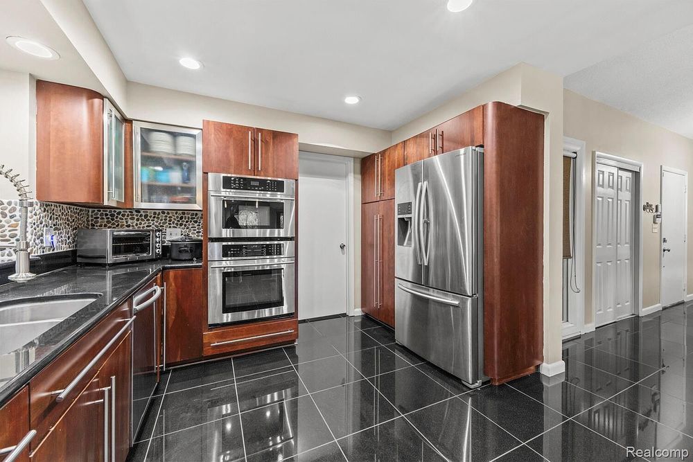 Cherry wood kitchen with stainless steel double oven, refrigerator, and black tile flooring.