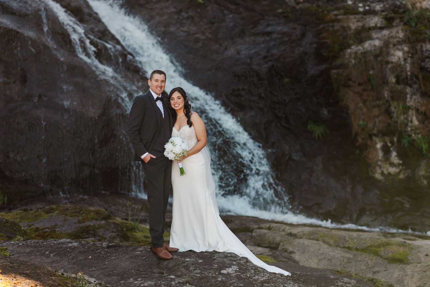 A couple in wedding attire poses in front of a cascading waterfall against rocky cliffs.