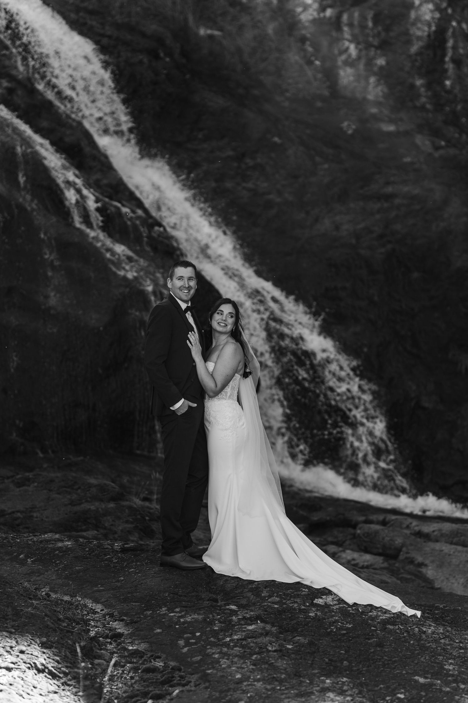 A black and white wedding photo series of a couple posing dramatically in front of a cascading waterfall.