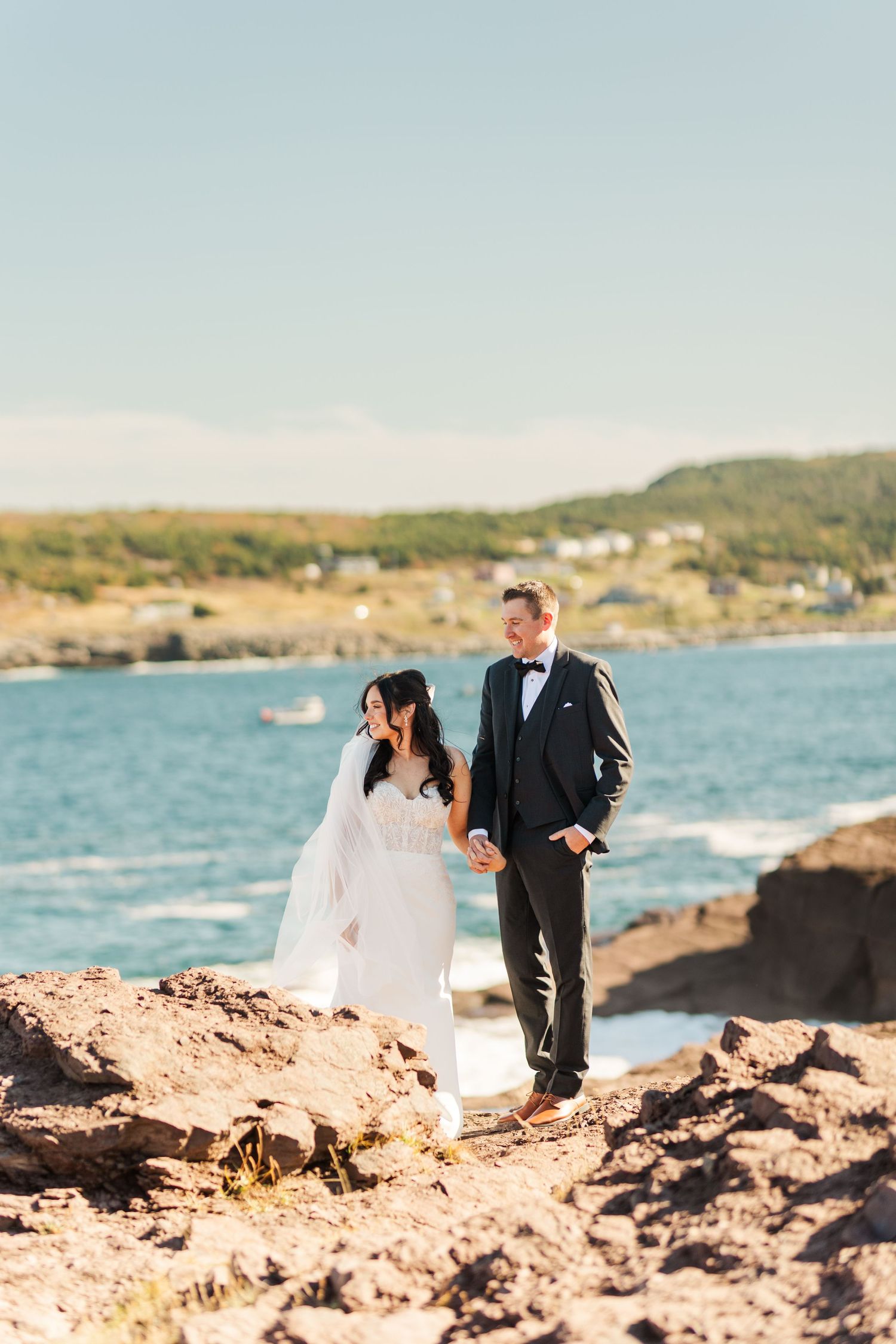 A couple in wedding attire walks along a rocky coastal shoreline with blue ocean waters and cliffs in the background.