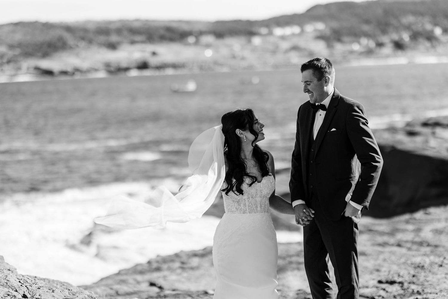 A couple in formal attire shares a romantic moment on a rocky beach shoreline in black and white.