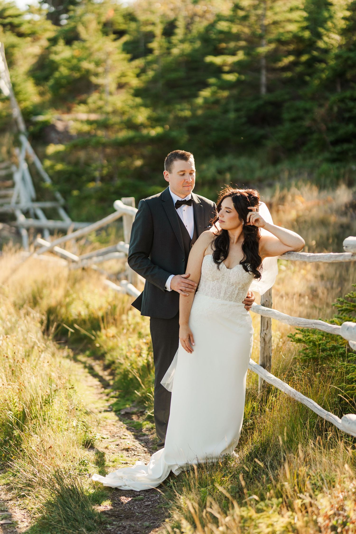 A couple in wedding attire poses along a rustic fence path surrounded by golden grass and evergreen forest.