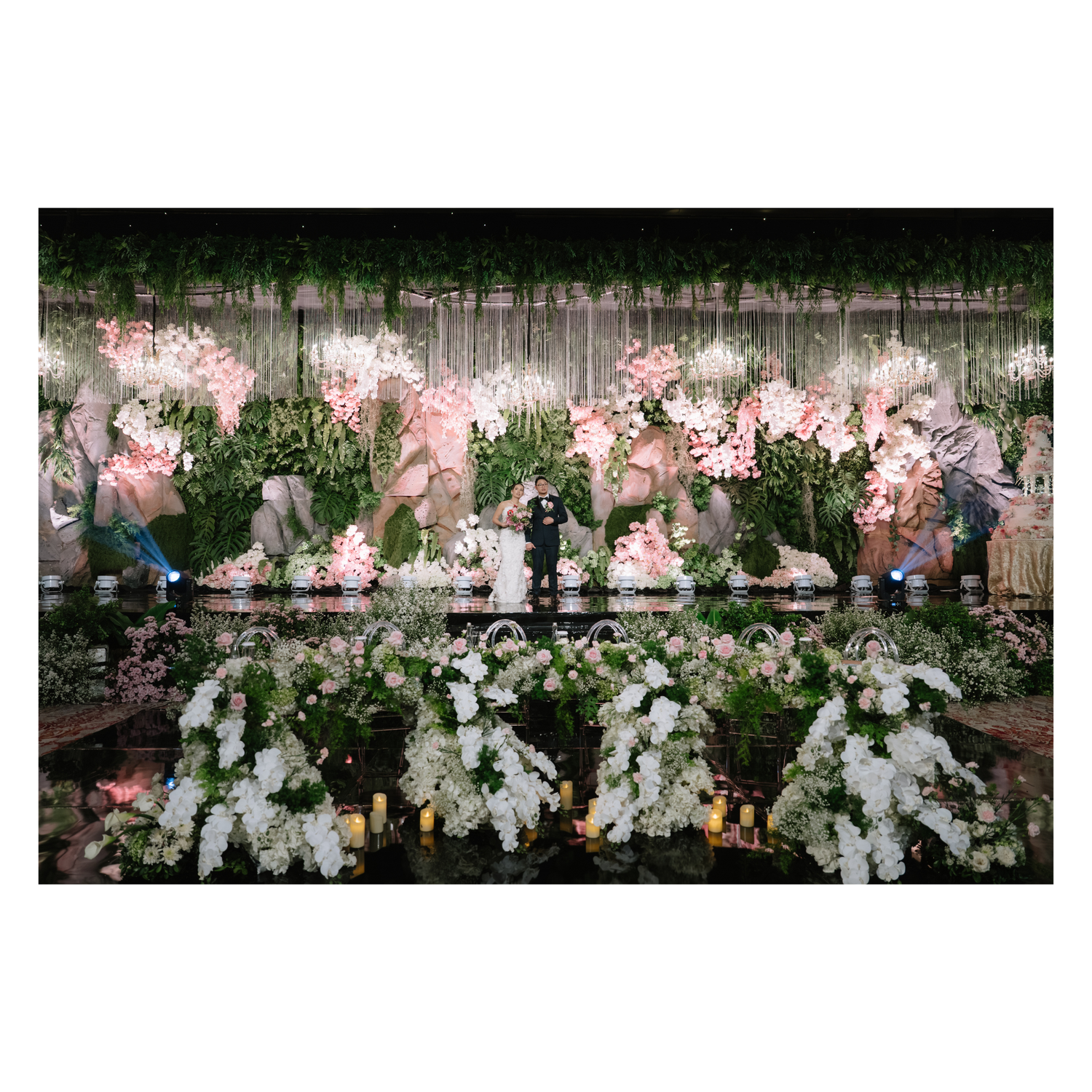 Elegant wedding reception stage with pink orchids, greenery backdrop, and white floral displays.