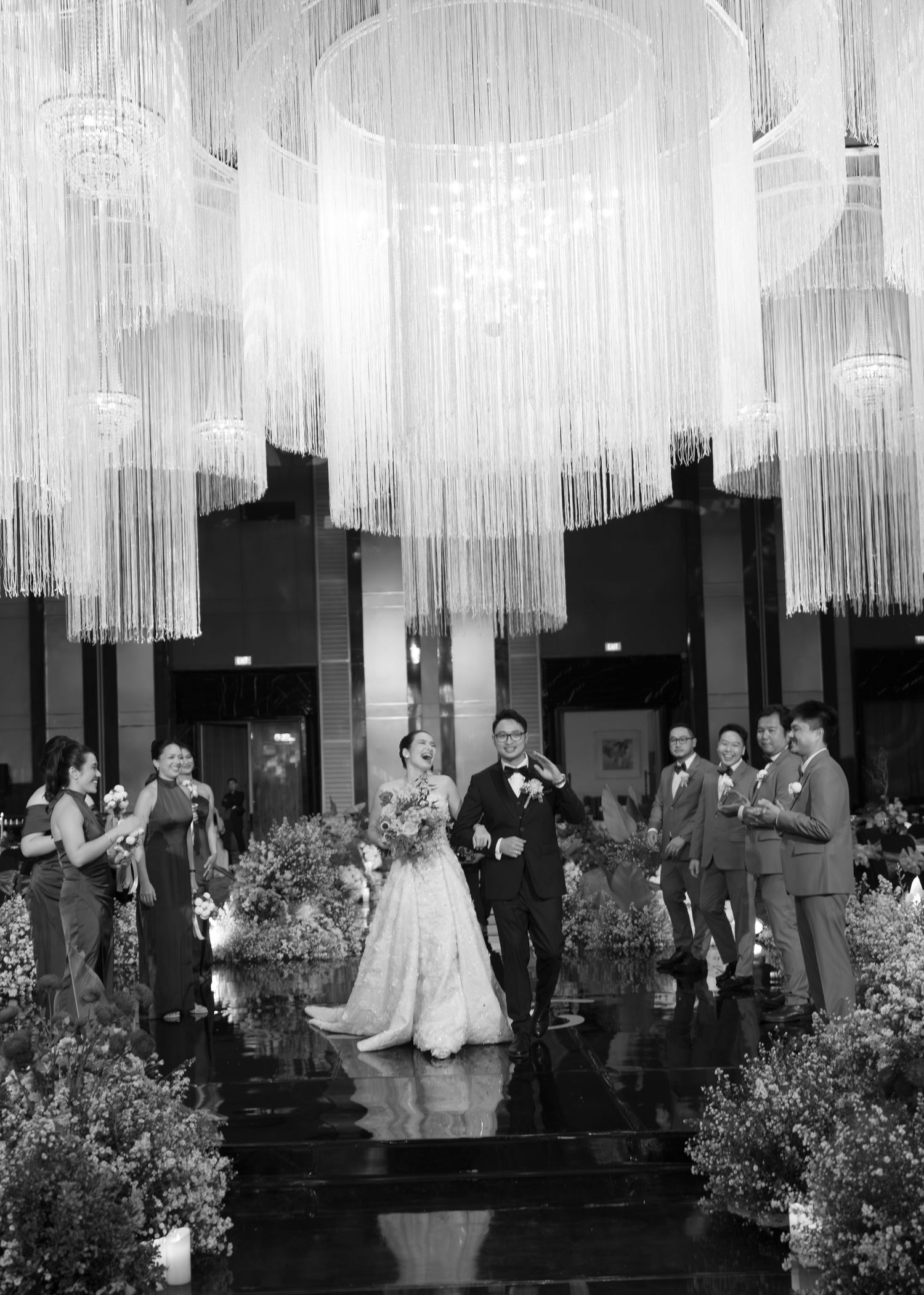 Black and white photo of bride and groom walking down aisle under stunning chandelier with wedding party.