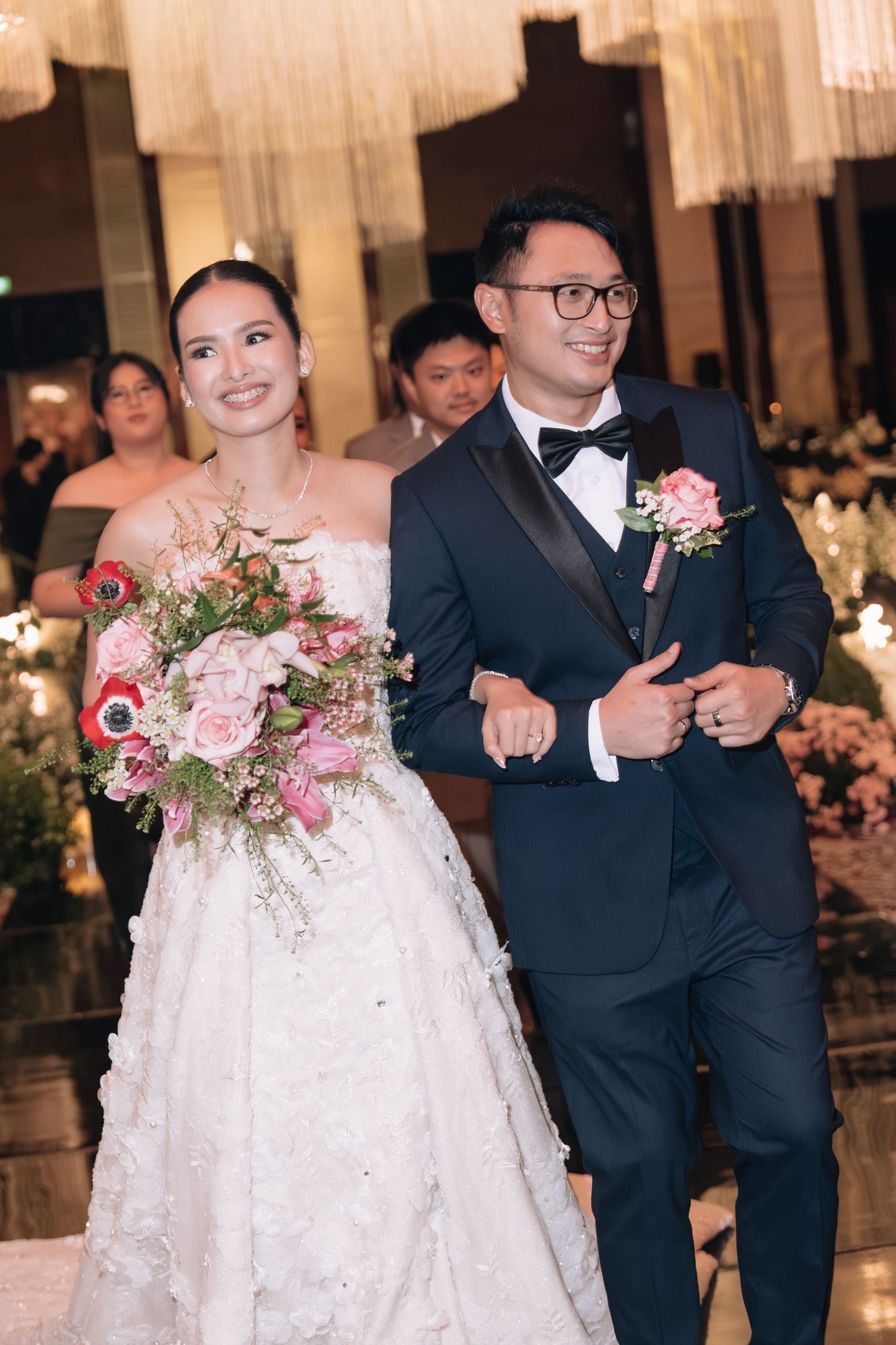 Smiling bride in white gown holding pink bouquet walks with groom in navy tuxedo at wedding reception.