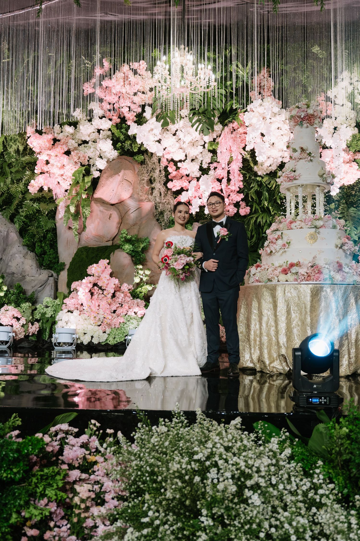 Bride and groom pose before elaborate pink floral backdrop with towering decorated wedding cake on stage.