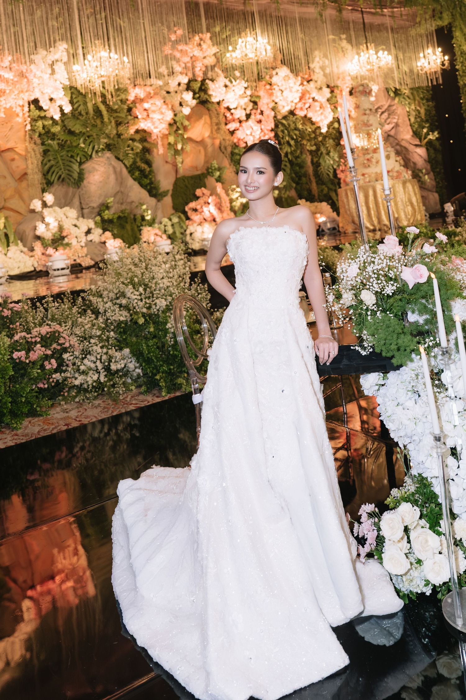 Elegant bride in strapless white gown with train poses before lush pink floral wedding reception backdrop.