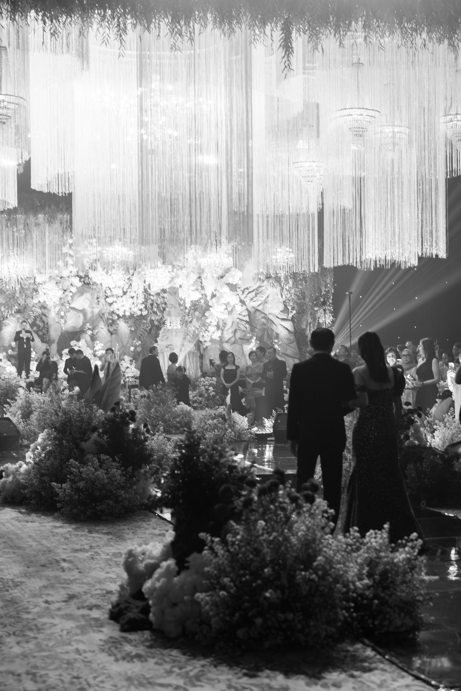 Black and white photo of crowded wedding reception with hanging crystal chandeliers and lush floral arrangements.