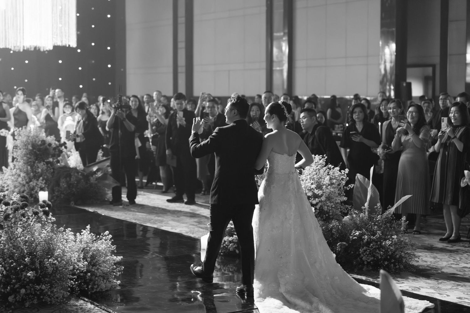 Black and white photo of bride and groom facing applauding wedding guests in grand reception venue.