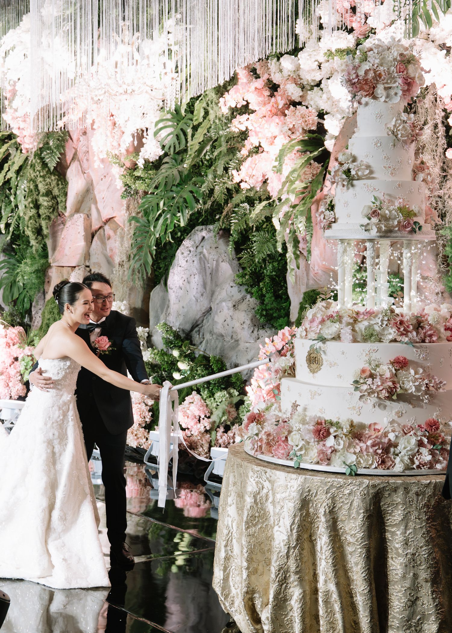 Bride and groom cut elaborate multi-tiered floral wedding cake adorned with pink blooms and greenery backdrop.