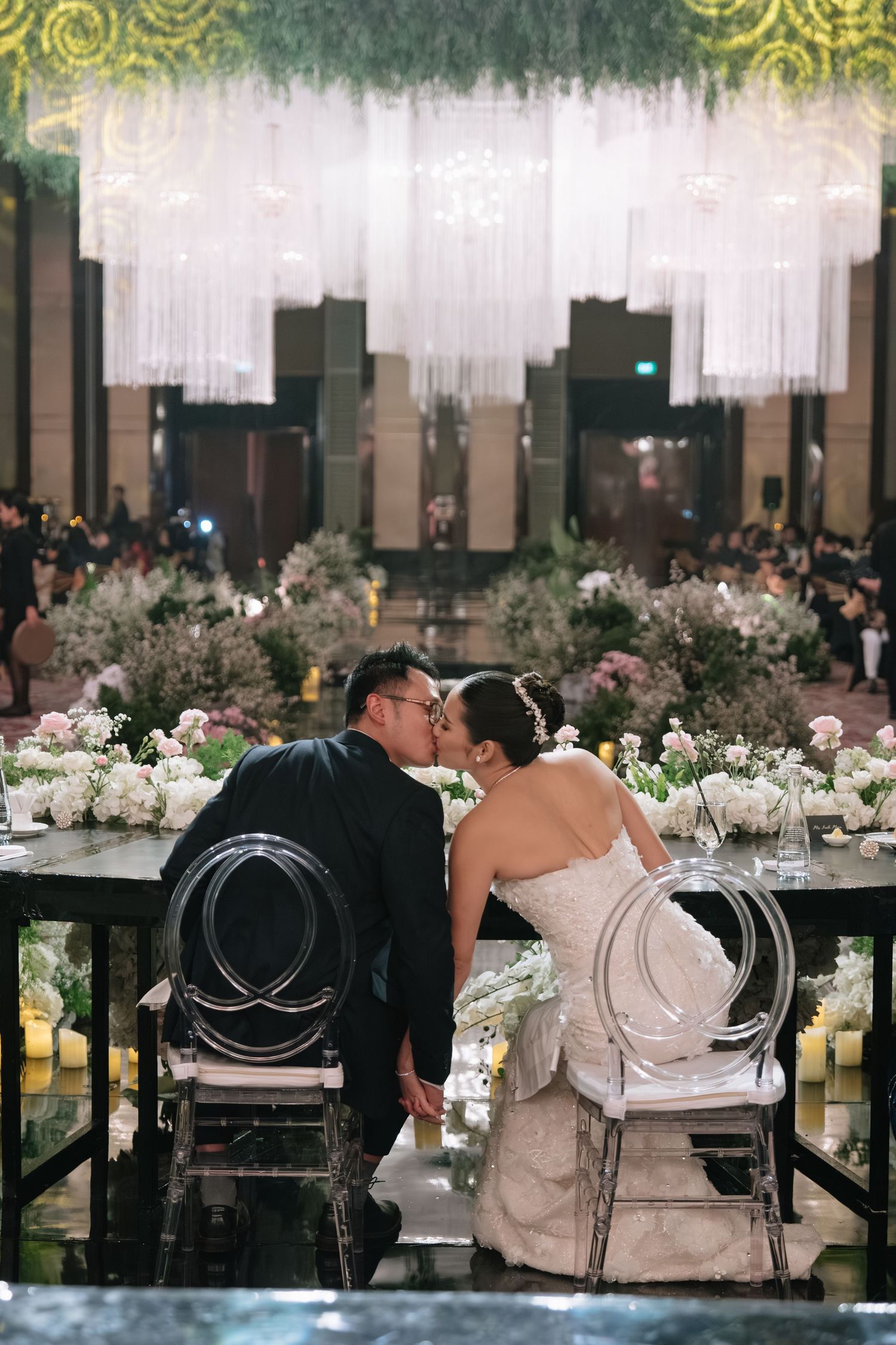 Bride and groom share a kiss at sweetheart table with white floral arrangements in elegant ballroom.