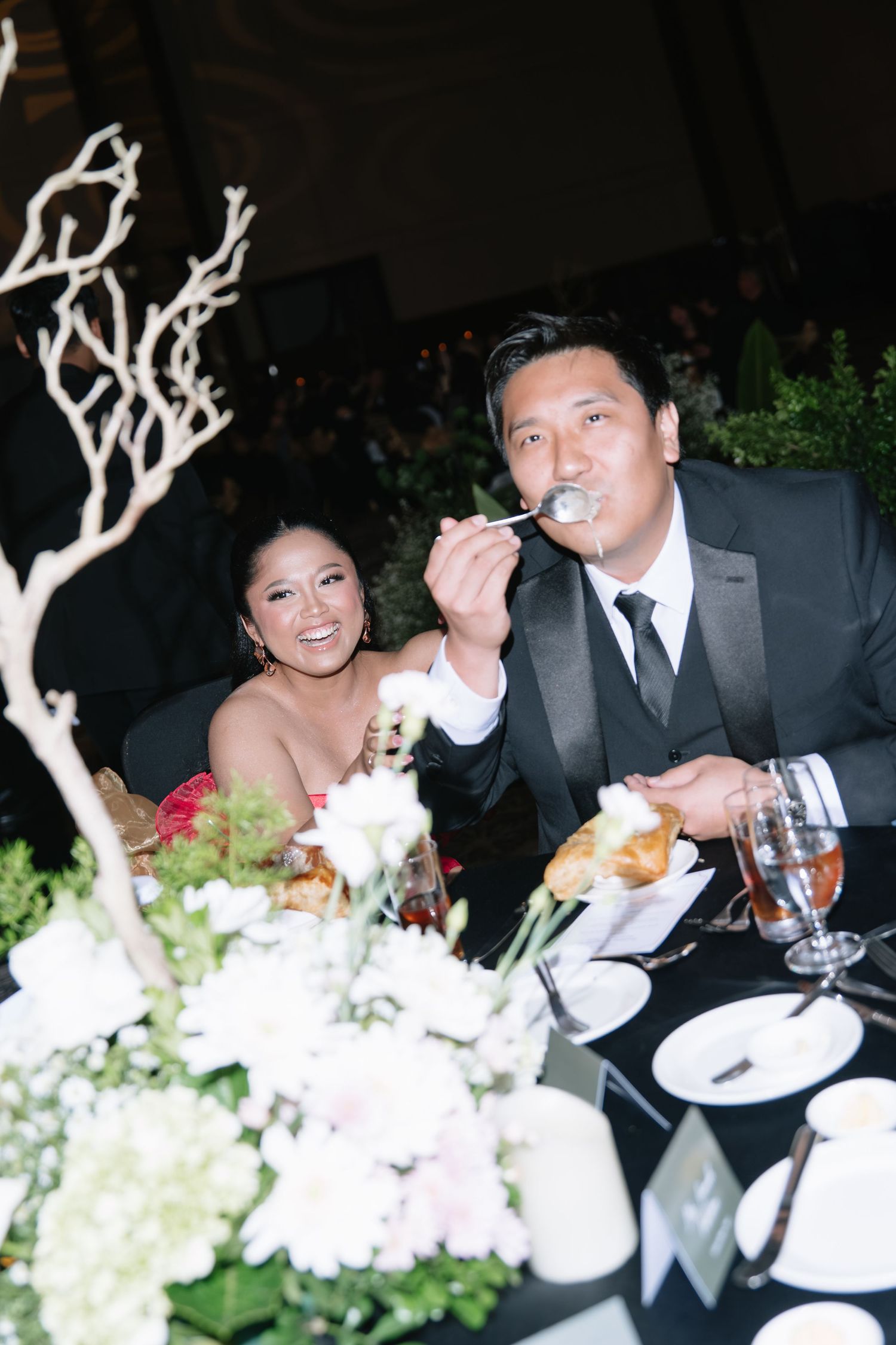Couple smiling at wedding reception dinner table with floral centerpiece and wine glasses.