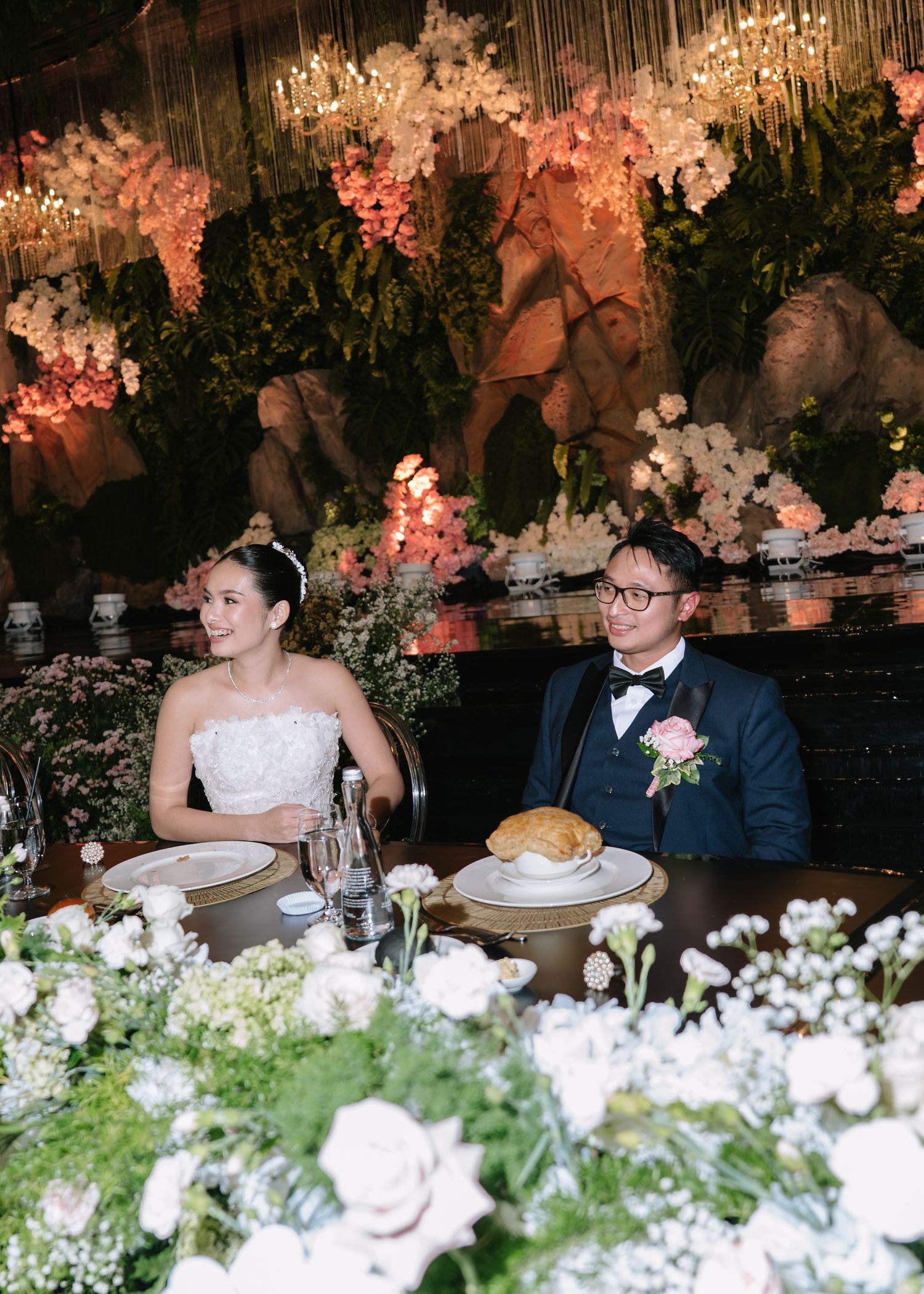 Bride and groom seated at elegant wedding reception table surrounded by lush floral arrangements.