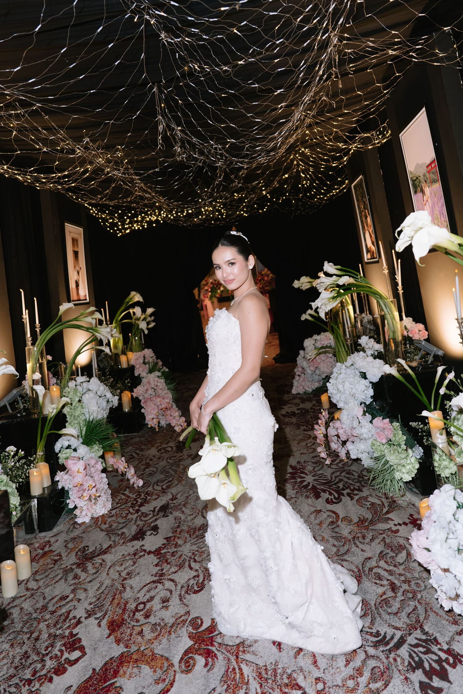 Bride in white lace mermaid gown holding calla lilies under twinkling fairy light canopy.