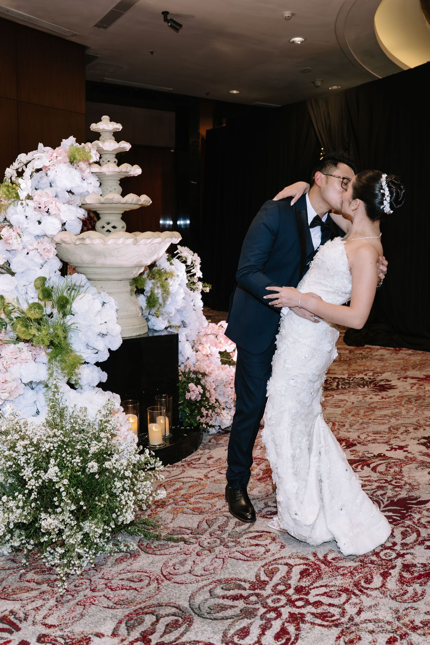 Bride and groom sharing romantic kiss near white floral fountain at wedding reception.