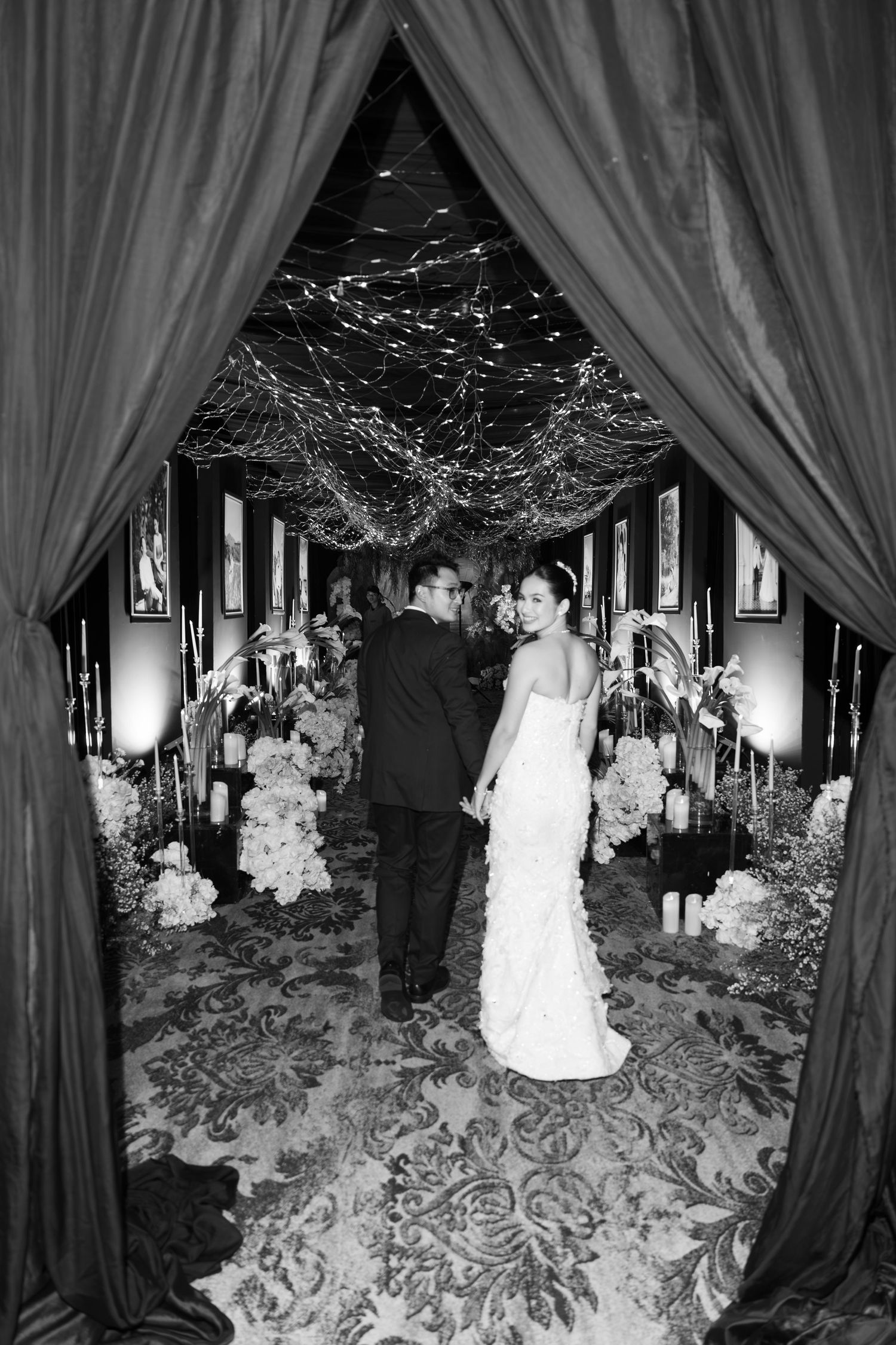 Black and white photo of bride and groom walking through draped floral wedding corridor.