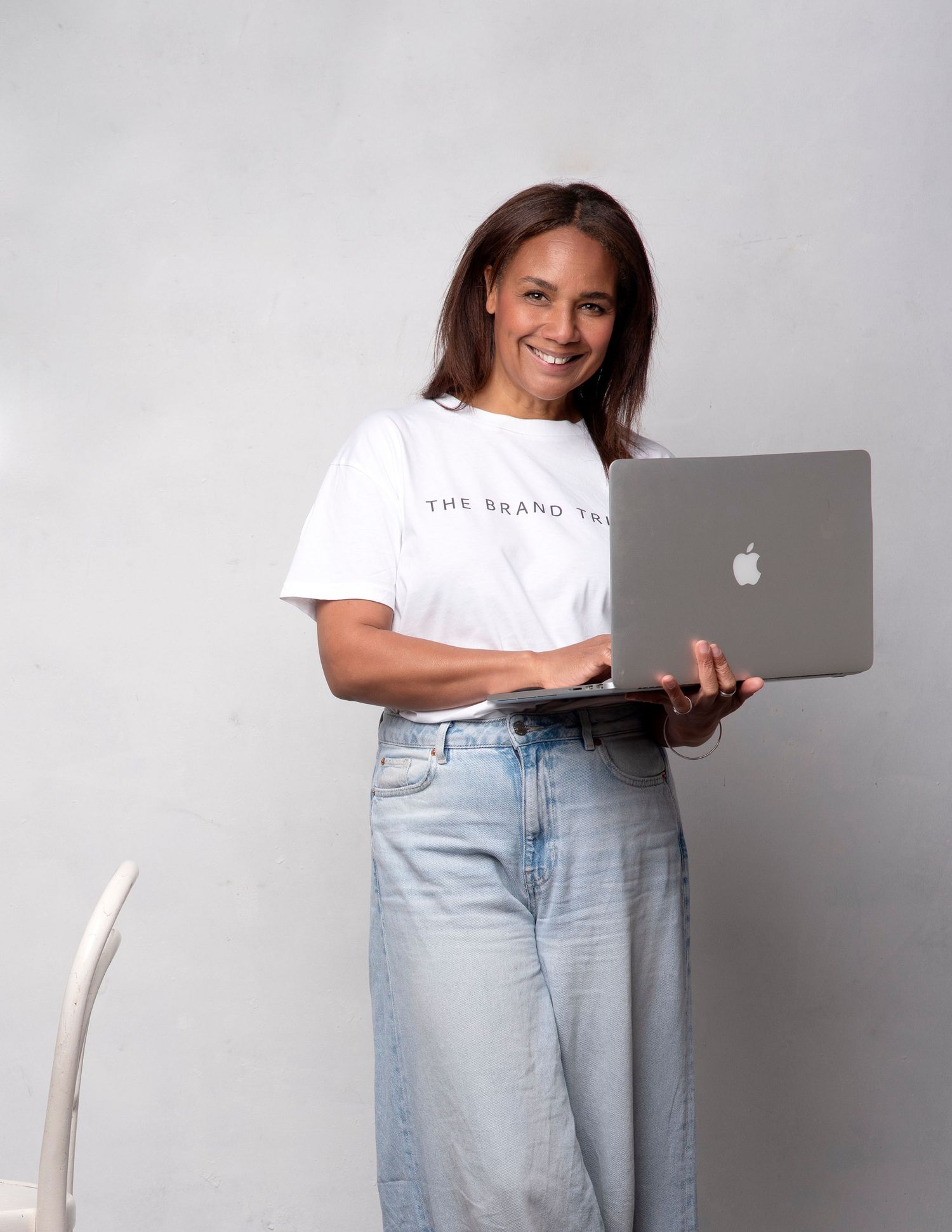 Person in white t-shirt and light jeans using MacBook laptop while sitting on chair against white background.