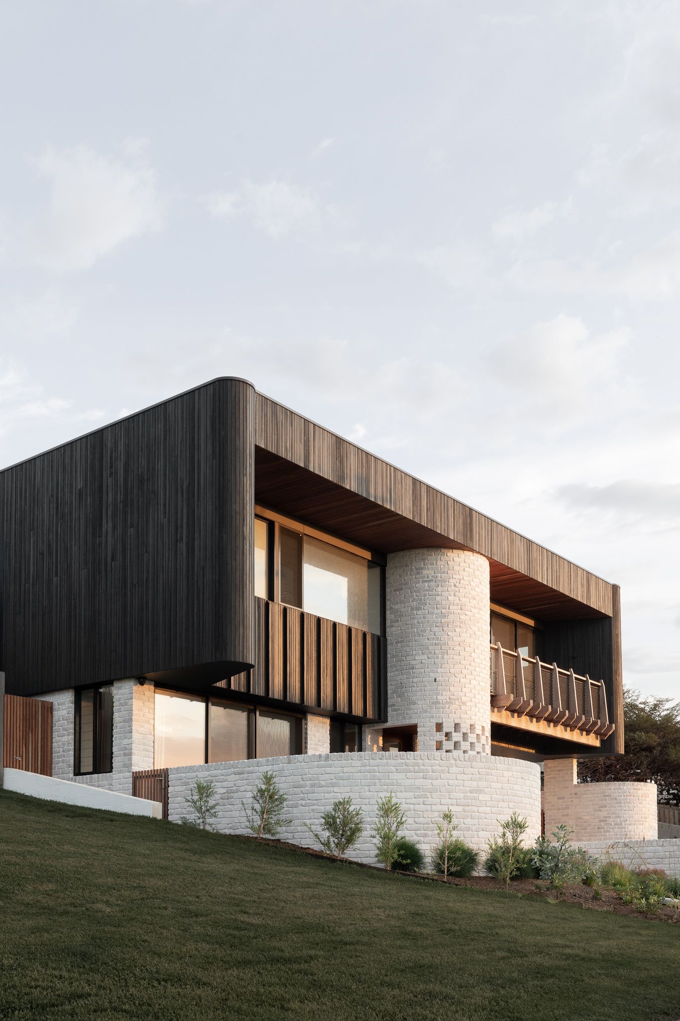 Modern hillside house with dark wood and white stone exterior features horizontal lines and large windows at dusk.