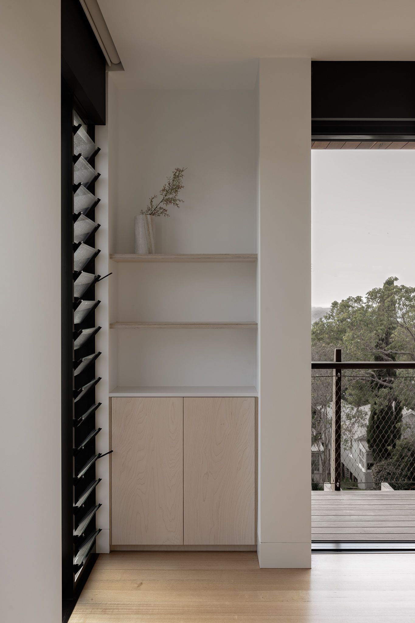 Modern minimalist interior with white floating shelves, wooden cabinet, and black louvered window panel beside a balcony view.
