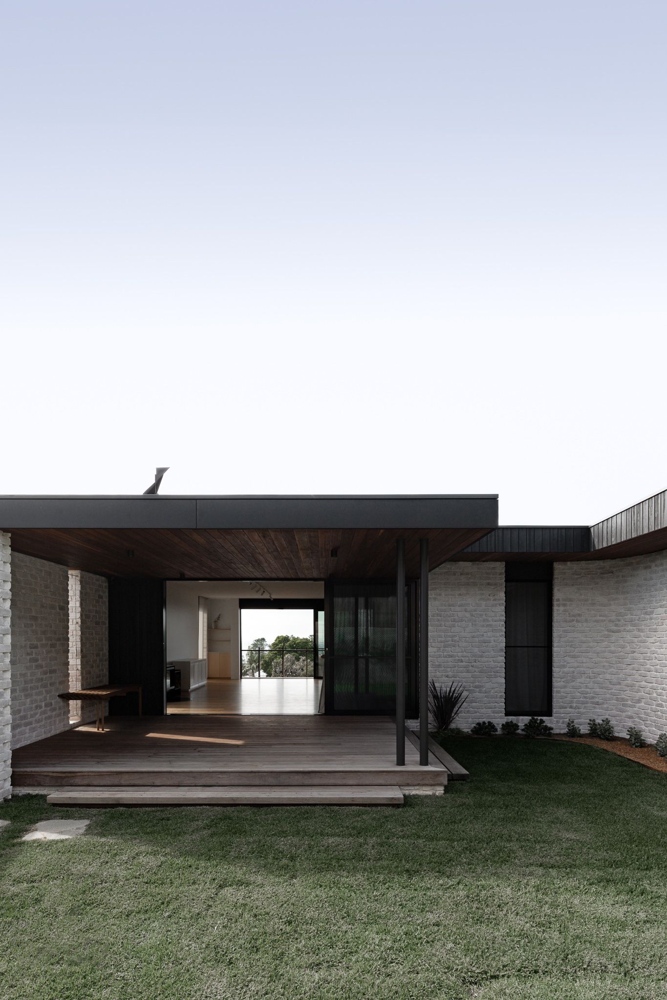 Modern minimalist house with dark brick exterior, wooden deck and large grassy lawn showcases clean architectural lines.