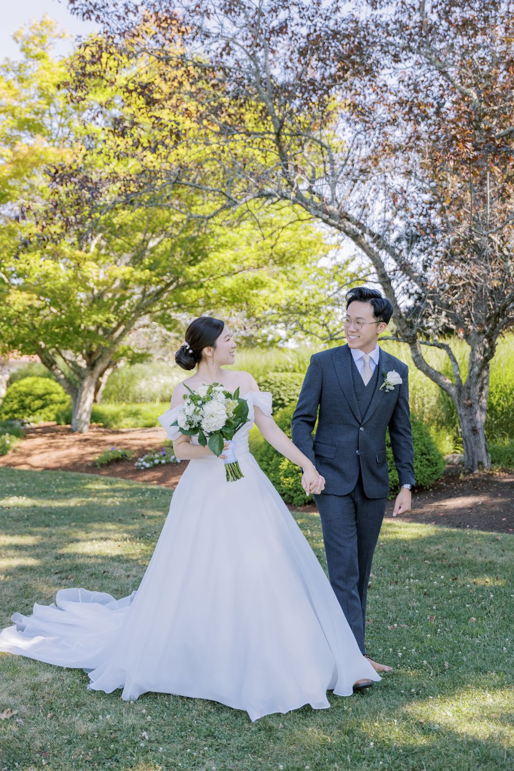 A wedding couple stands together on a lush garden lawn with blooming trees in the background.
