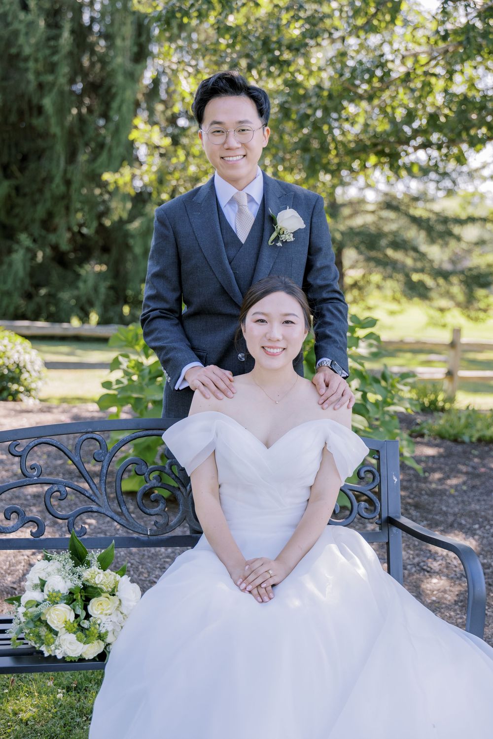 A wedding couple poses on an ornate garden bench surrounded by greenery and flowers.
