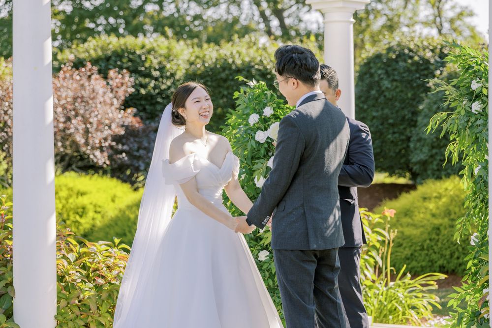 A couple shares a joyful moment under a white garden pergola surrounded by lush greenery on their wedding day.