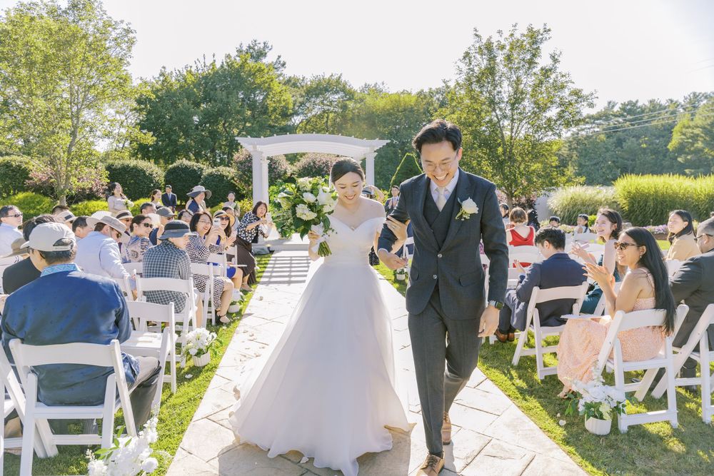 A scenic outdoor wedding ceremony takes place under a white gazebo with guests seated on white chairs on a sunny day.
