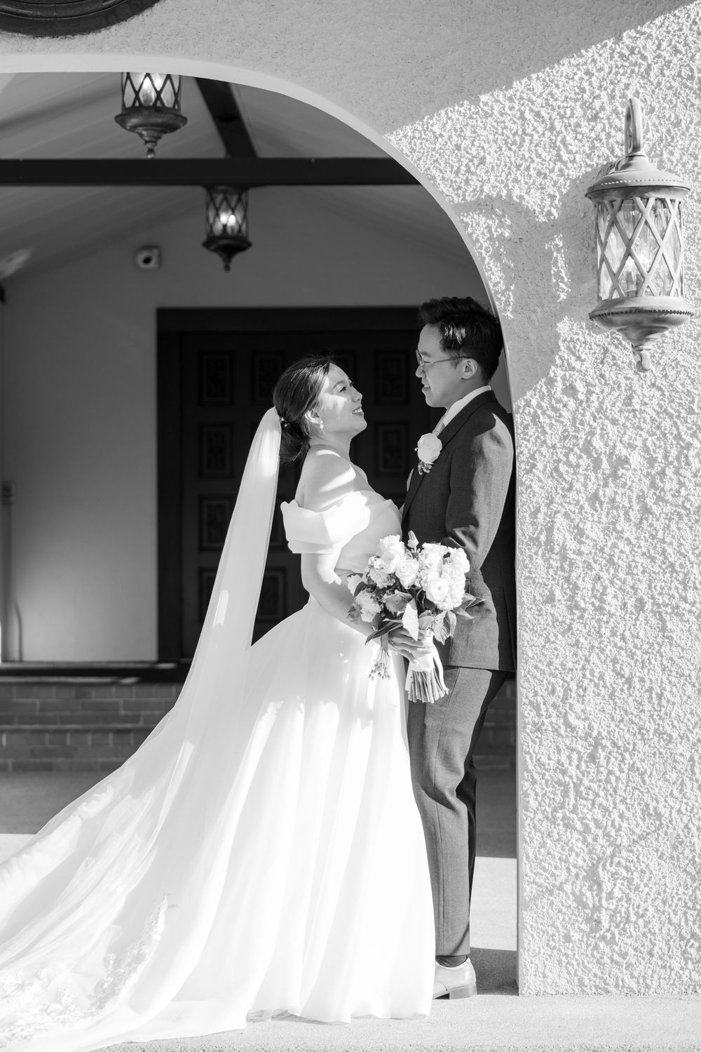 A romantic black and white wedding photo of a couple in formal attire standing under an archway with hanging lanterns.